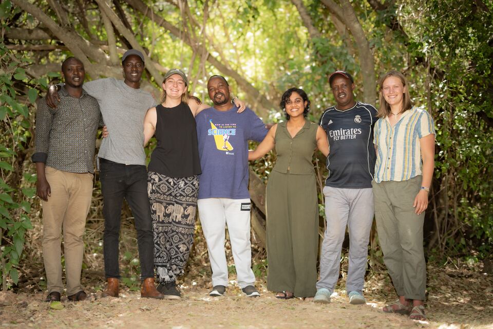 Seven members of the Talek field team standing arm in arm beneath the trees at camp
